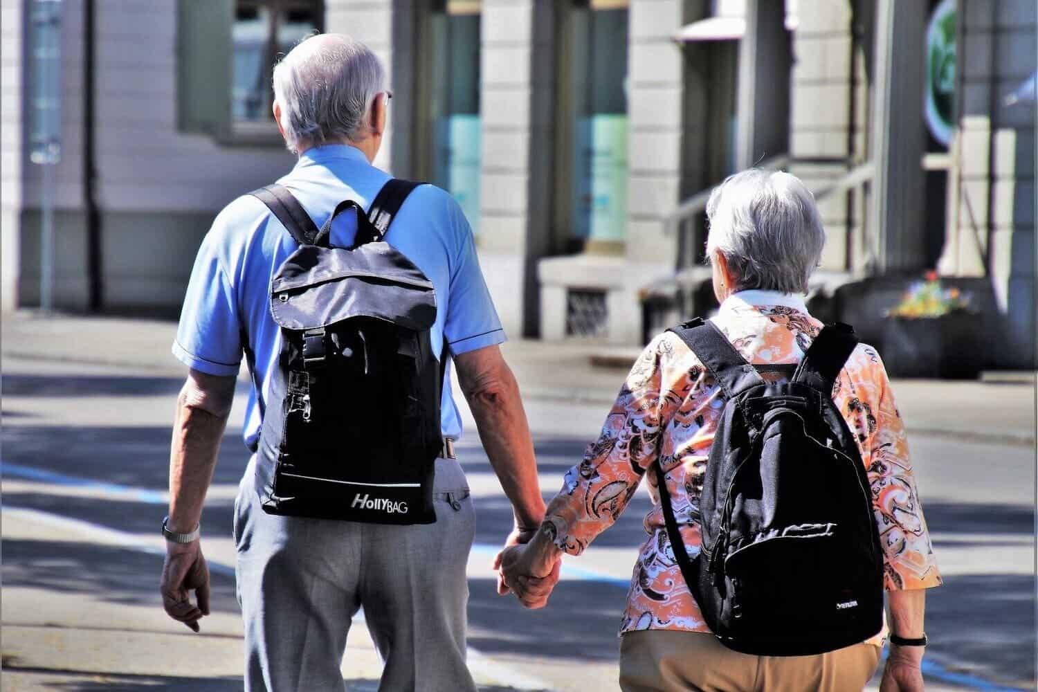 una pareja de adultos mayores, van caminando por la calle agarrados de manos y cargando mochilas pesadas