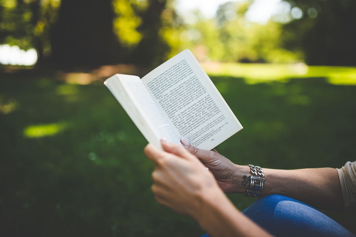 las manos de una persona joven que sostiene un libro abierto, mientras está sentado(a) en un espacio lleno de naturaleza