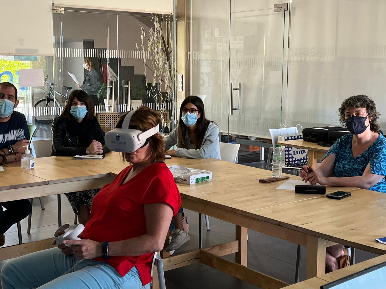 la imagen fue tomada en la reunión con cuidados paliativos, en donde cinco personas reunidas en una sala, en primer plano, una señora usando gafas de Realidad Virtual, al fondeo, cuatro personas con mascarilla viendo lo que sucede
