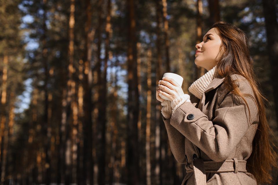 una mujer en un bosque, en sus manos lleva una taza de café, sonríe mientras ve hacia el cielo