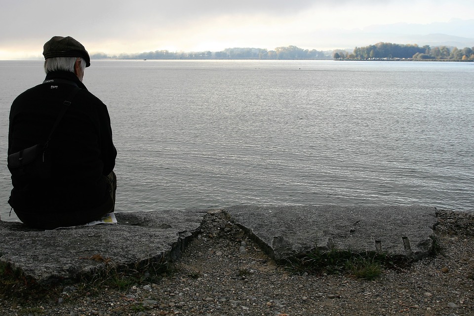 un hombre mayor, sentado en el borde de un muro, viendo hacia el mar que tiene en frente