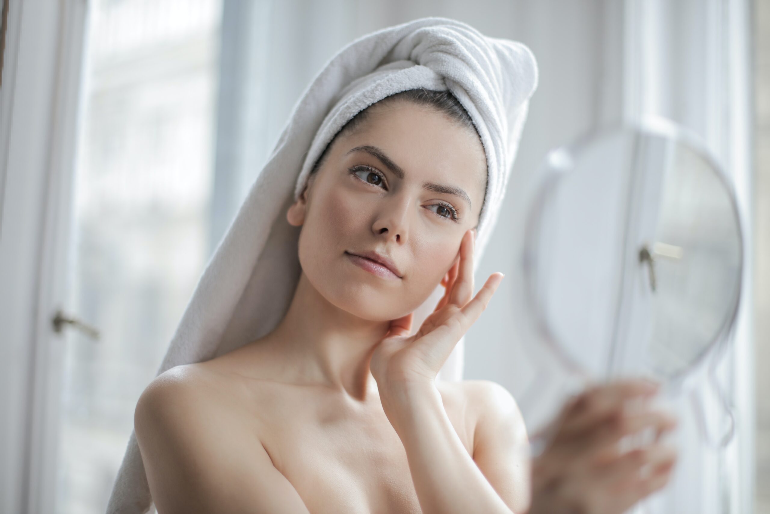 una mujer que se esta viendo en un espejo, con su cabello envuelto en una toalla, mientras se va haciendo masajes en la cara