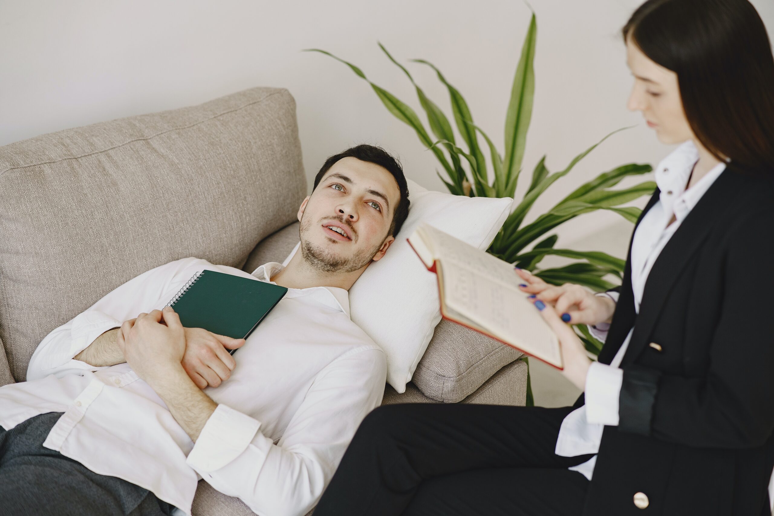 un hombre tumbado en un sofa con una libreta en us mano, mientras hace sesión con una terapeuta que va tomando notas