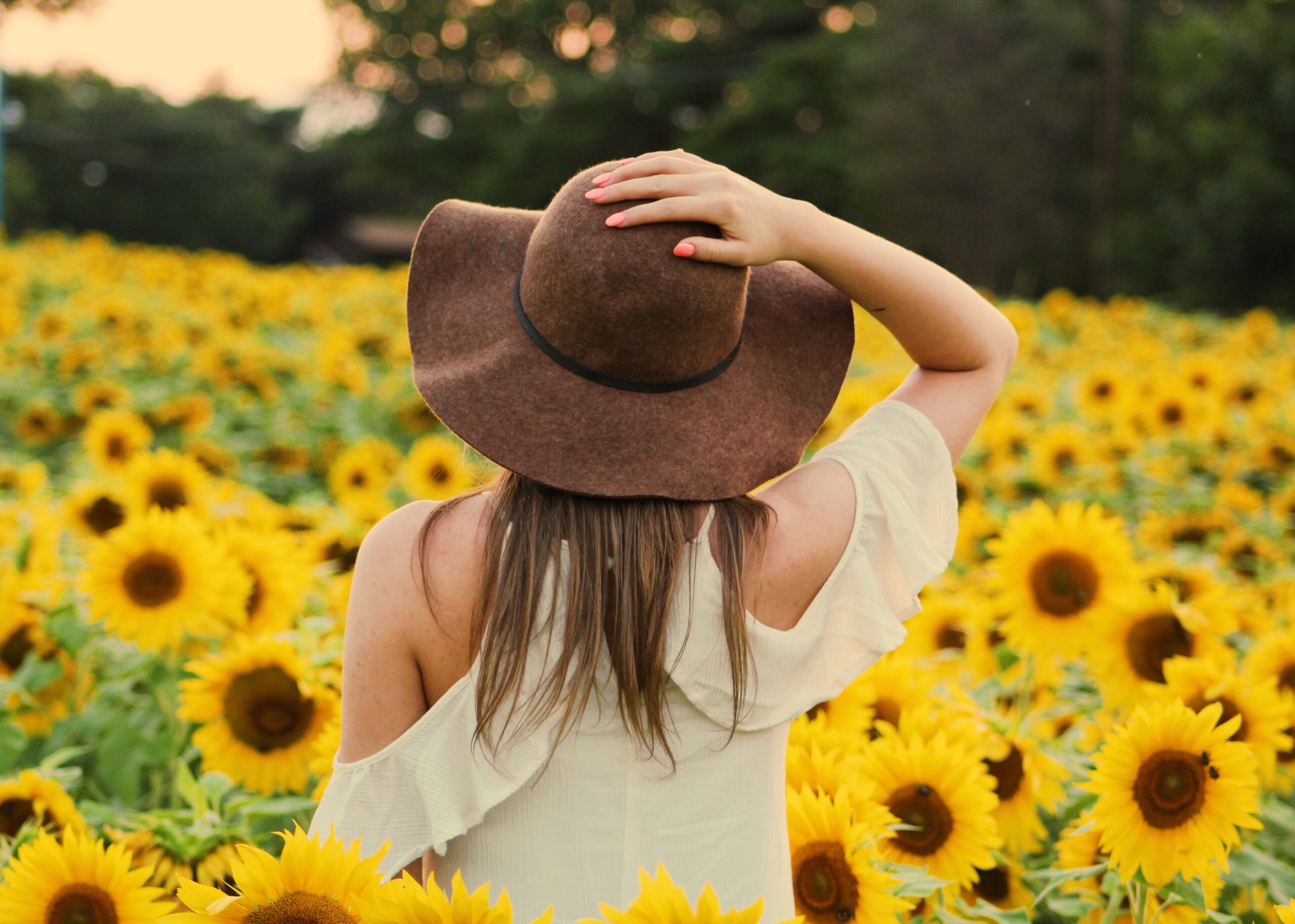 una mujer se agarra el sombrero que lleva en la cabeza, mientras admira la naturaleza que tiene en frente, un campo lleno de girasoles