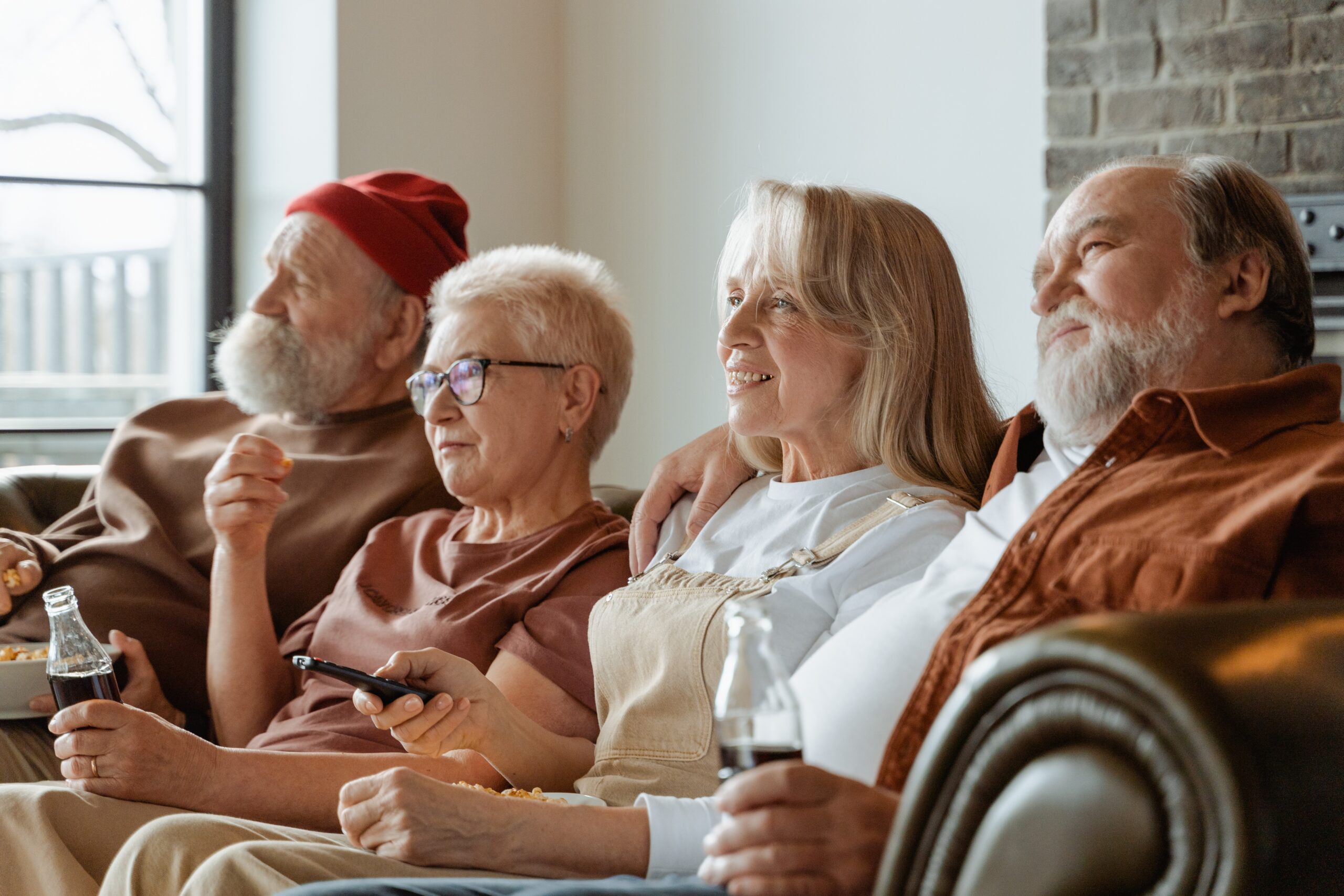 cuatro personas mayores, sentadas en un sofá, comiendo palomitas y bebiendo refrescos mientras ven la televisión