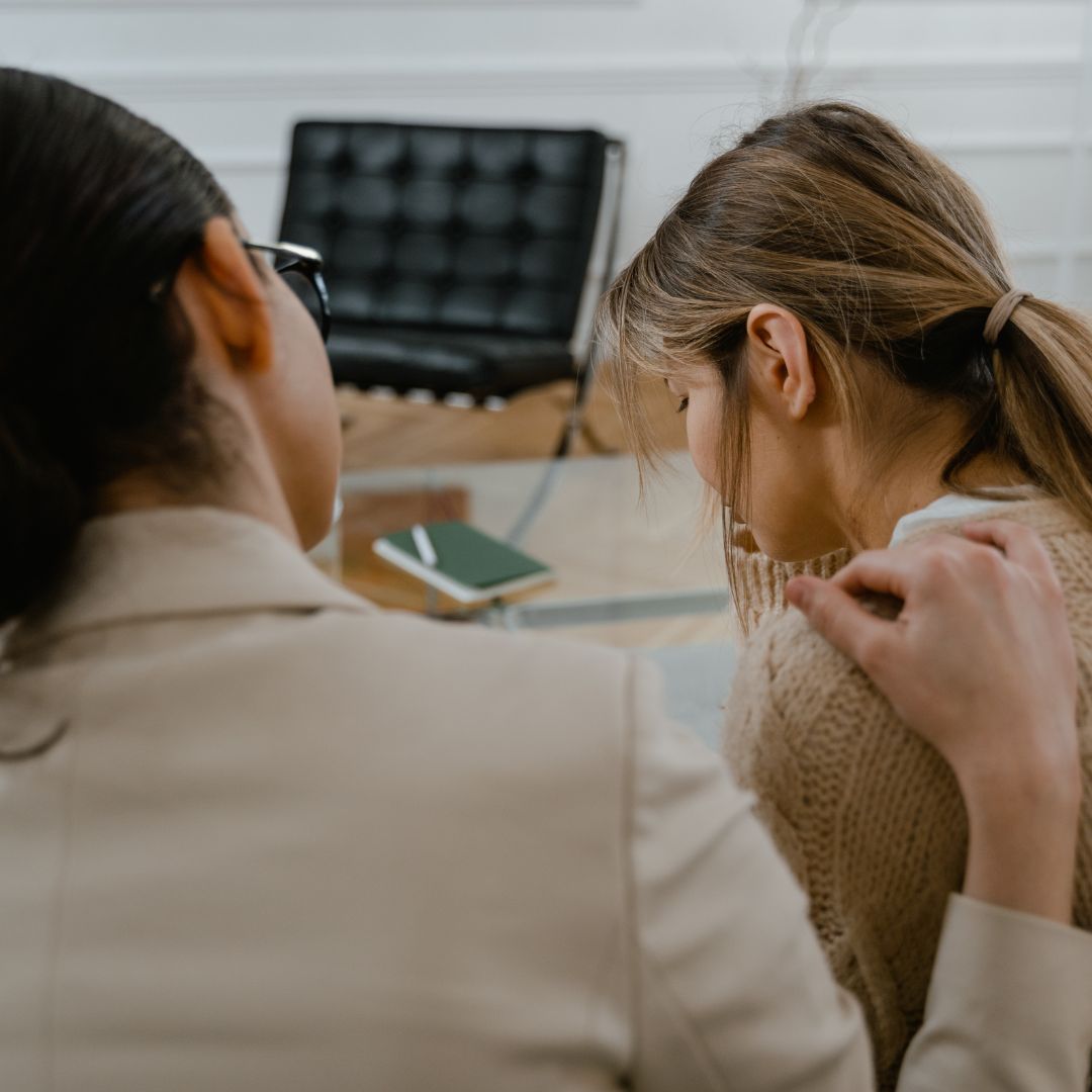 una mujer que aparenta estar triste o preocupada, mirando hacia el suelo mientras que otra mujer que aparenta ser su terapeuta o medico, tiene su mano sobre el hombreo de la primera, la imagen hace referencia a la importancia del apoyo emocional a las personas que padecen la enfermedad de pick