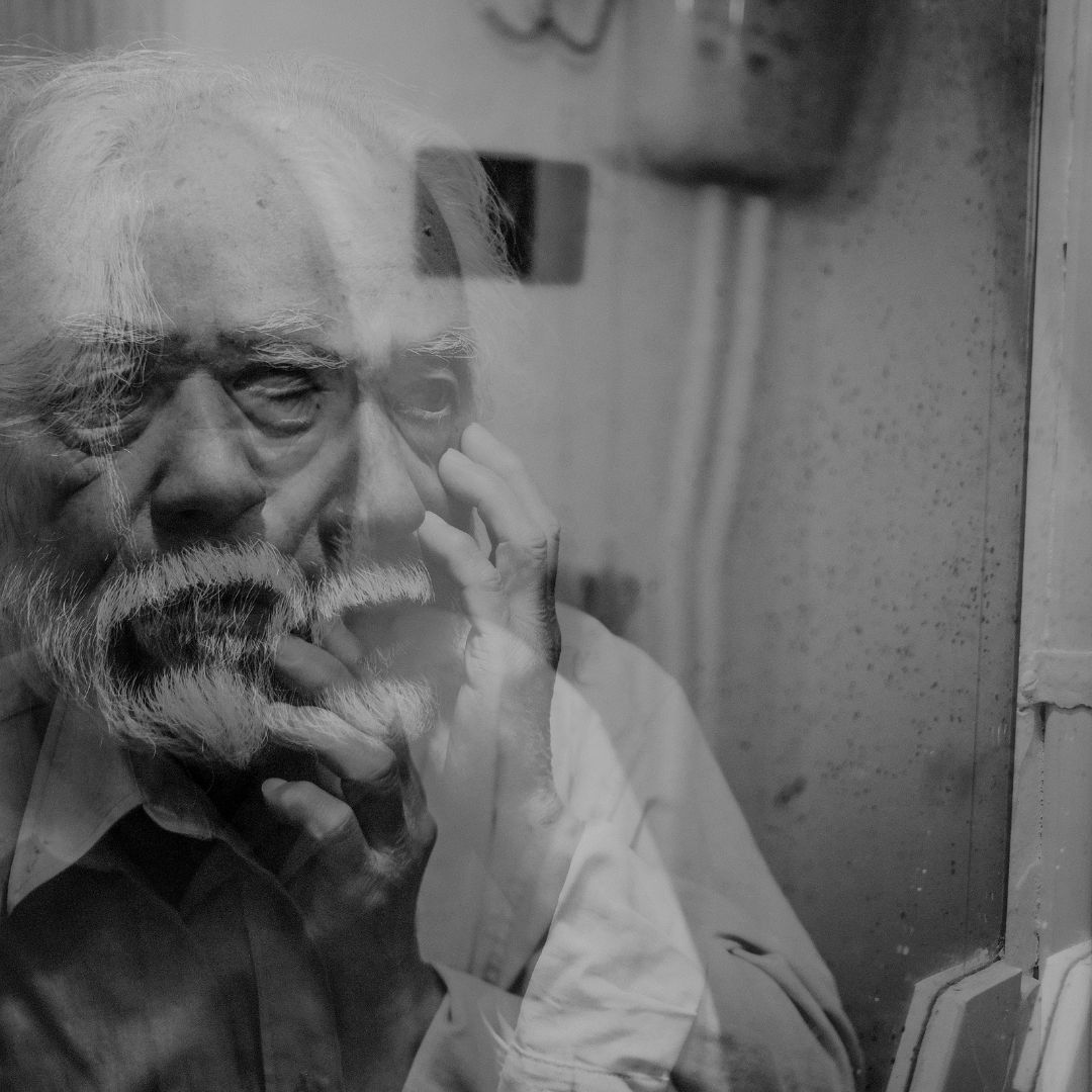 en la imagen a blanco y negro, se ve a un hombre mayor que se ve en un espejo de manera borrosa, lo que el hombre ve es su rostro preocupado . la imagen hace referencia a los síntomas de la demencia