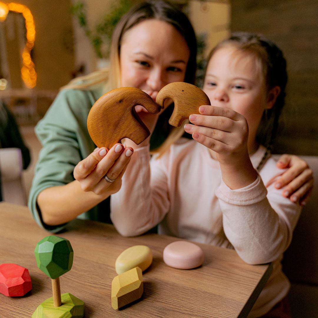 en la imagen se ve una mujer y una niña, jugando y/o haciendo ejercicios cognitivos. Mientras juegas, la mujer tiene un brazo alrededor de la niña y su otra mano sostiene una de las manos de la niña. La imagen hace alusión a una niña con discapacidad intelectual