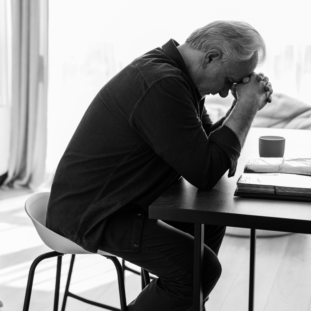 imagen a blanco y negro de un hombre que esta sentado y tiene sus brazos apoyados en una mesa, mientras sus manos sostienen su cabeza.La imagen proyecta soledad y tristeza, síntomas de soledad no deseada