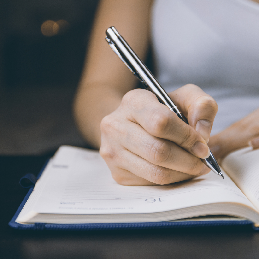 en la imagen se logra identificar la mano de una mujer que esta escribiendo con un bolígrafo en una agenda, haciendo referencia a la dificultad que algunas personas pueden tener para escribir debido a alguna lesión cerebral, es decir, agrafia