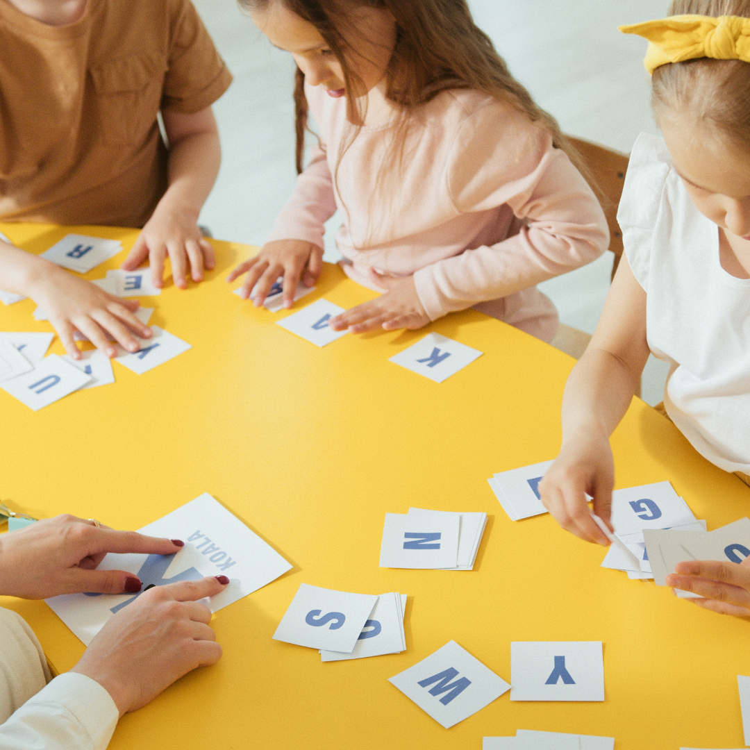 en la imagen se logra ver 3 niños en lo que parece ser un aula, en donde hay una profesora enseñándoles el abecedario o enseñándoles a crear palabras (en una mesa tienen recortes de letras, cada niño tiene distintas letras y la maestra les esta enseñando un recorte en concreto, en el cual se aprecia la letra k y de bajo una palabra), 2 de los niños están concentrados en sus letras, mientras que un tercero, parece estar viendo las letras de su compañera. Con esta imagen se hace referencia a que los problemas de aprendizaje suelen aparecer en la infancia