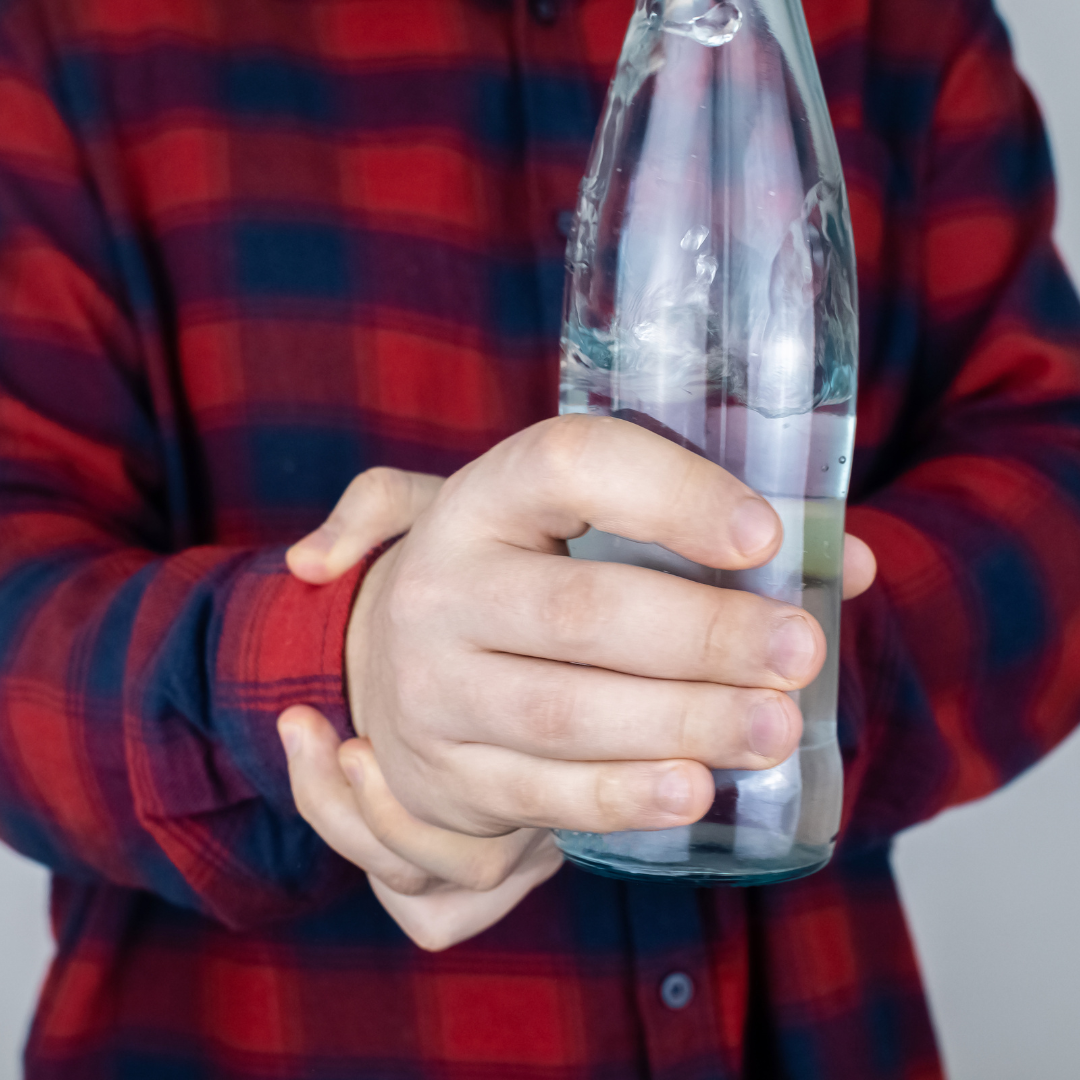 en la imagen se logra ver el torso de un hombre; con su brazo derecho flexionado hacia a delante, en su mano respectiva mano sostiene una botella de agua, mientras que la mano izquierda agarra con fuerza la muñeca derecha. Esto hace referencia a uno de los síntomas de la distonía