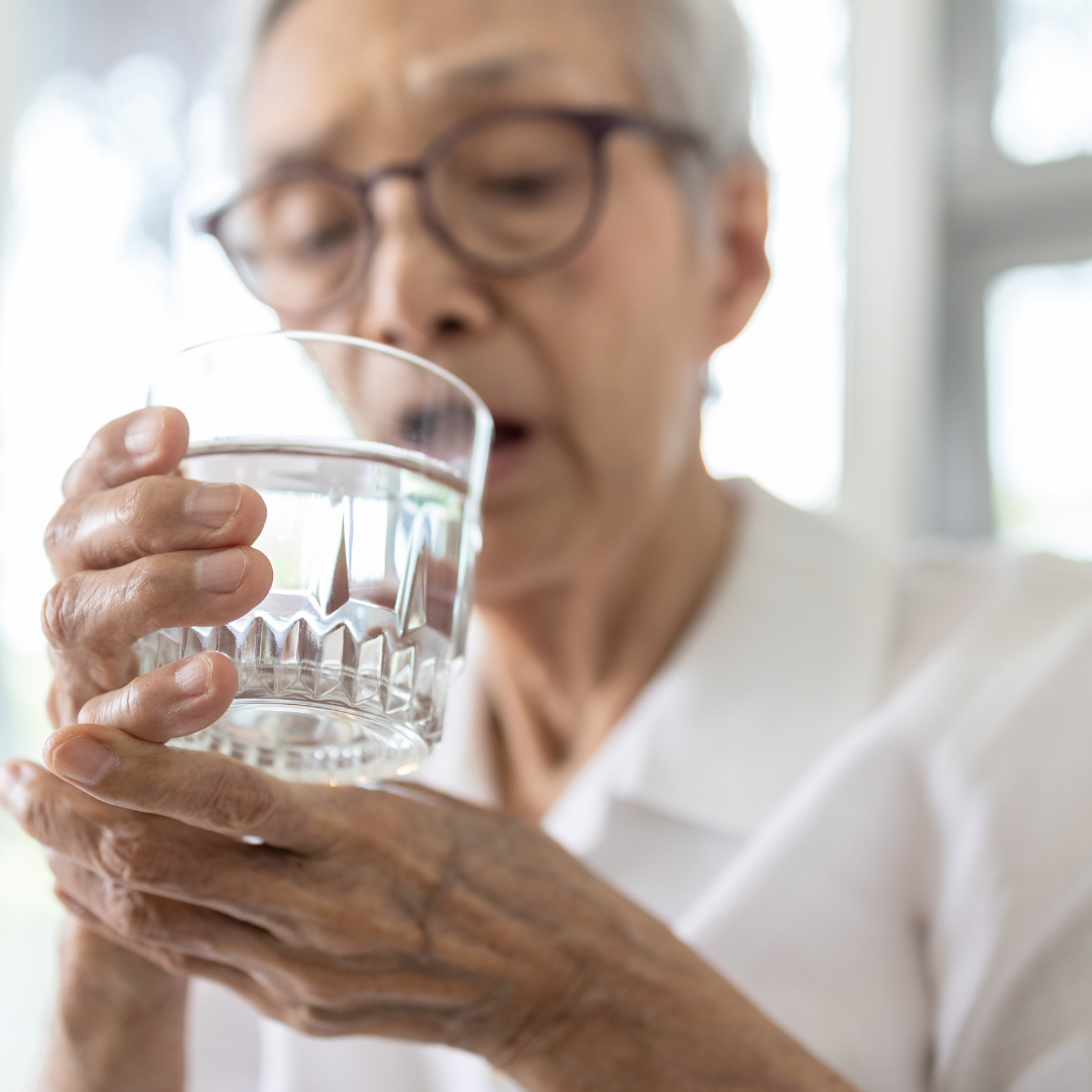 una persona mayor mientras hace ademan de beber agua, sostiene una de sus muñecas debido a que sufre de temblor esencial, lo que puede hacer que derrame lo que contiene el vaso que sostiene