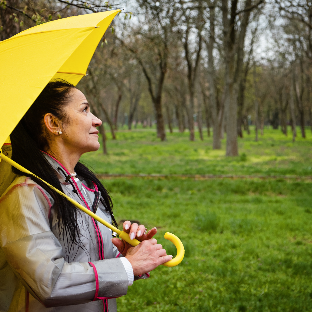 una mujer en un bosque, mira hacia el cielo mientras parece sonreír, aunque la foto es grisácea porque parece que lloverá, la mujer sostiene un paraguas amarillo. Imagen que refleja la resiliencia en pacientes con enfermedades neurológicas